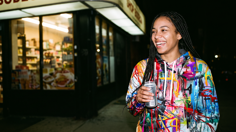 A woman laughing outside a corner store at night in the Paint Riot hoodie
